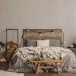Stylish grey chair with blanket and log of wood next to warm double bed with wooden headboard and light bulbs